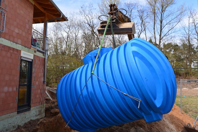 Ein Bagger hievt einen großen Erdtank in ein ausgehobenes Loch vor einem Neubau.