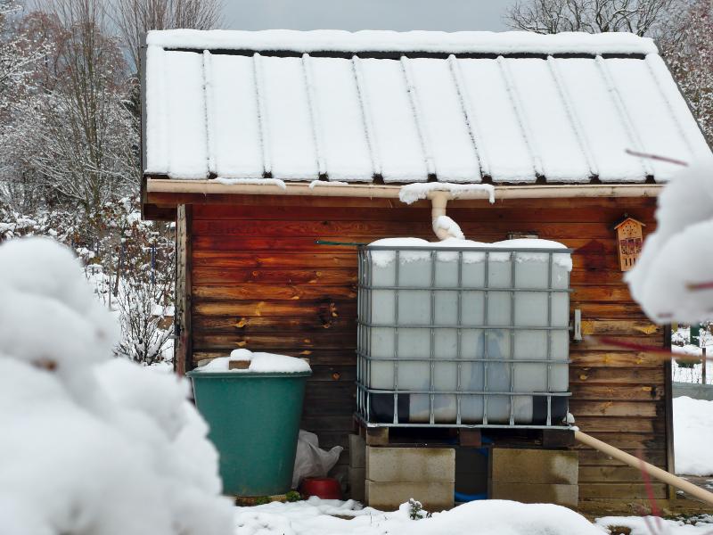 Vor einer tiefverschneiten Gartenhütte ist ein IBC-Container platziert, der an das Fallrohr angeschlossen ist. Regenwasserbehälter