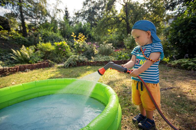 Ein kleiner Junge füllt mit einem Schlauch der an eine Wasseranschlussdose angeschlossen ist ein Plantschbecken