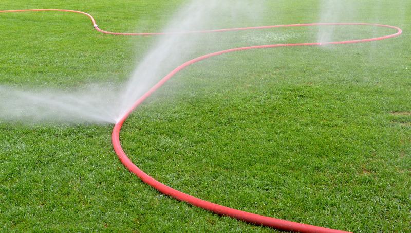 Aus einem Sprühschlauch kommen hohe Wasserfontänen die als Nebel auf eine Rasenfläche niedergehen